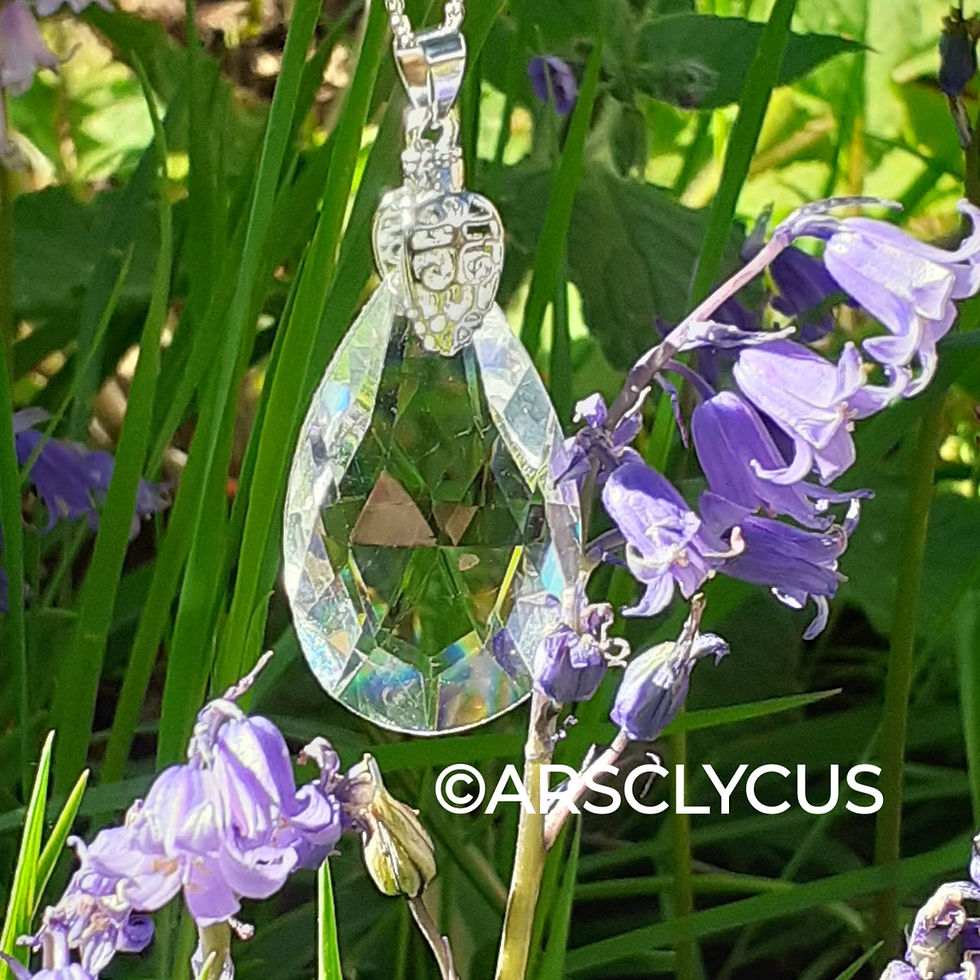 A crystal pendant hangs among purple flowers, reflecting light. The background is lush and green. ©ARPSCLYCUS is visible.