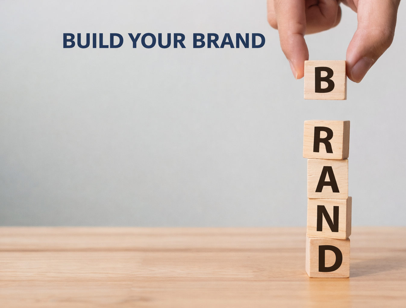 Hand stacking letter blocks spelling BRAND on wooden surface, with "BUILD YOUR BRAND" text in the background. Neutral backdrop.