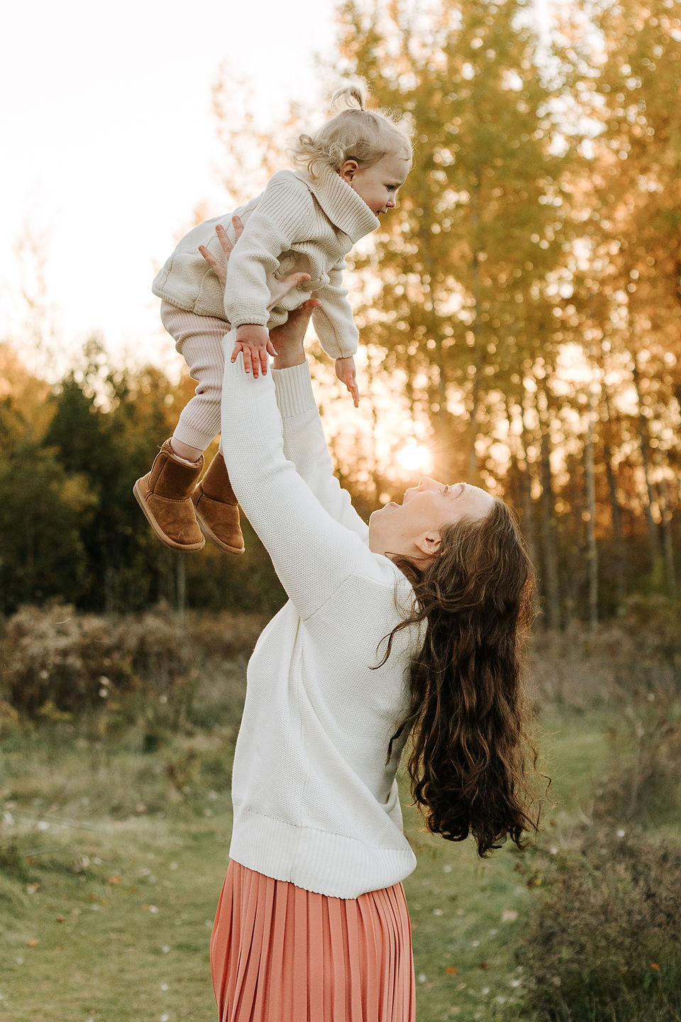 A woman lifts a young girl in the air on a grassy path under trees in the fall