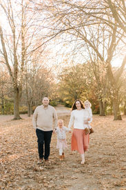 A man and woman walk hand in hand with a little girl while the woman has another little girl in her arms and they walk on a path under trees in the fall