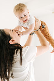 A woman in a white t shirt holds a one year old boy in the air and smiles at him