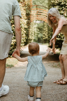 A one year old girl holds hands with an older man and reaches her hand out to an older woman who looks down at her and smiles as she reaches her hand out