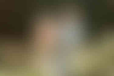 An older man holds a one year old girl and smiles as an older woman stands next to them and rests her hand on the girl as they stand amongst flowers