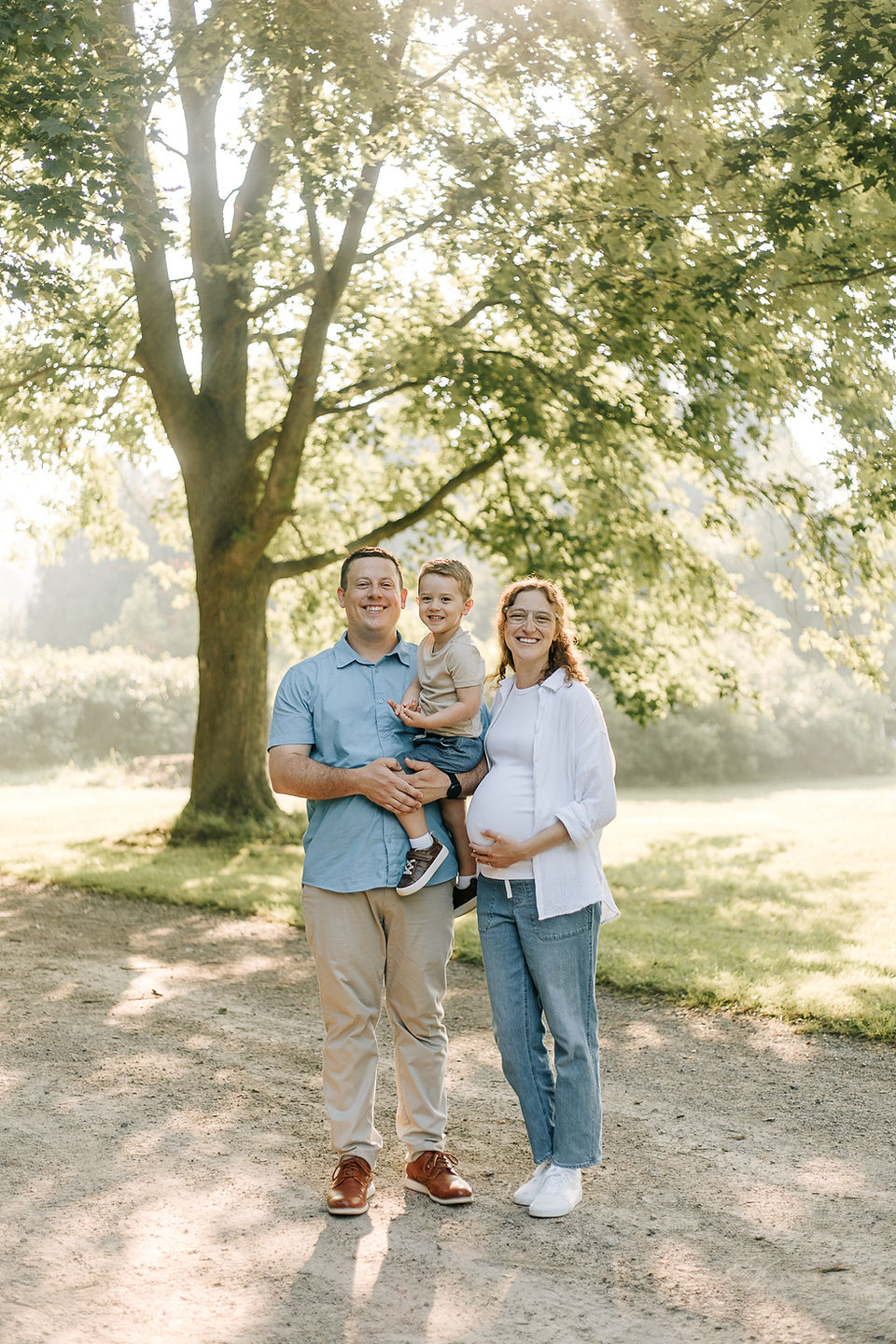 A man and pregnant woman hold a young boy on a dirt path under trees