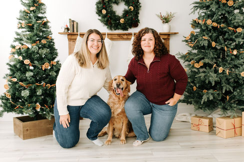 Two woman kneel in between two Christmas trees as a golden retreiver dog sits between the woman