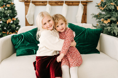 Two girls hug as they sit on a couch in between two Christmas trees