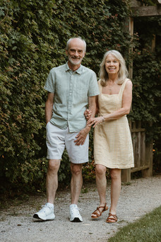 An older woman and man hold hands and walk beside a tall hedge