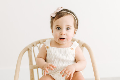 A one year old girl sits on a small rattan chair