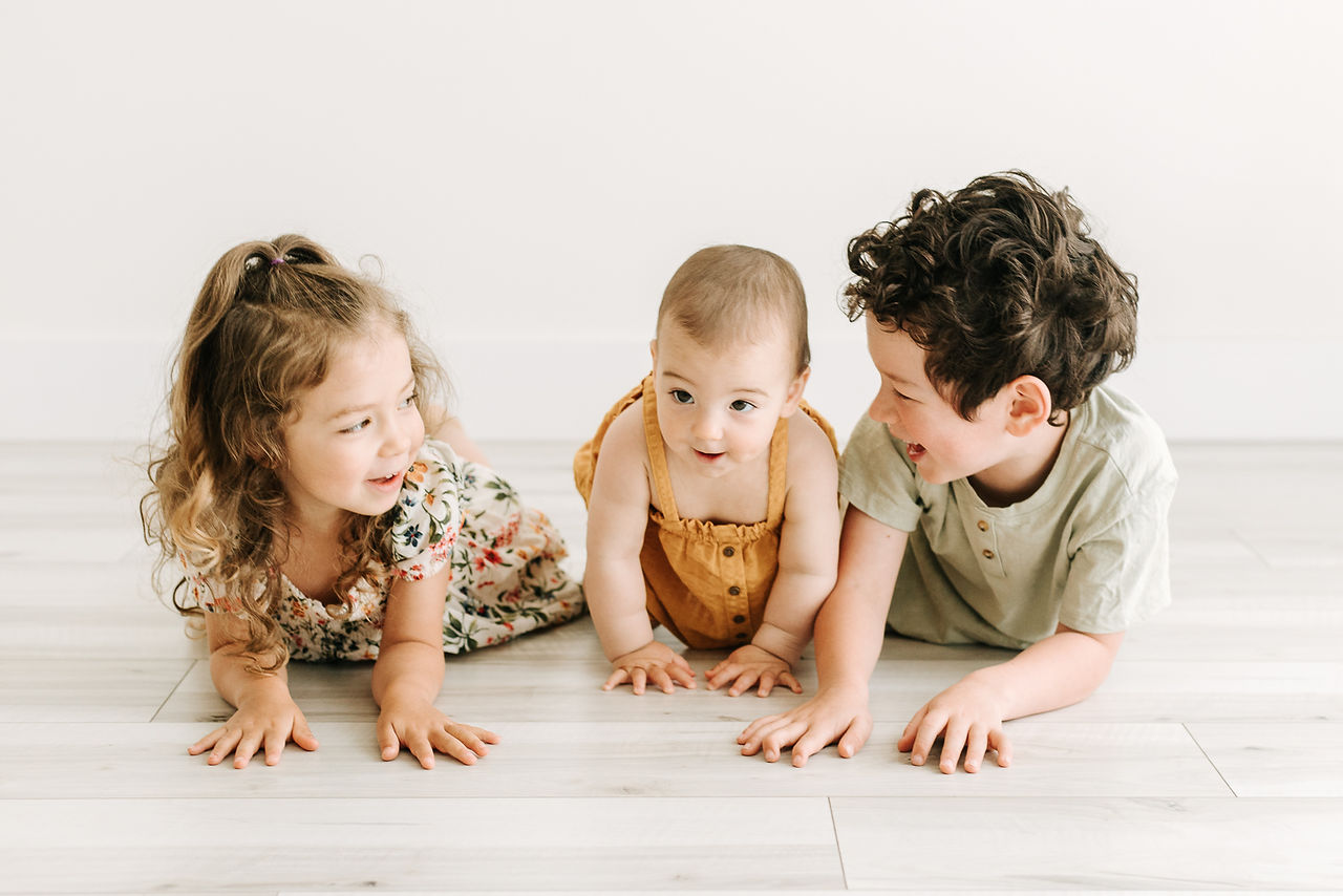 A baby girl, young girl, and young boy lie on their stomachs on the floor and look at each other