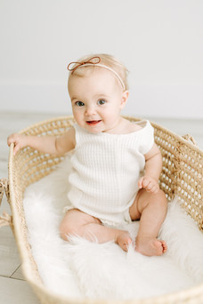 A one year old in a white outfit and hair bow sits in a moses basket