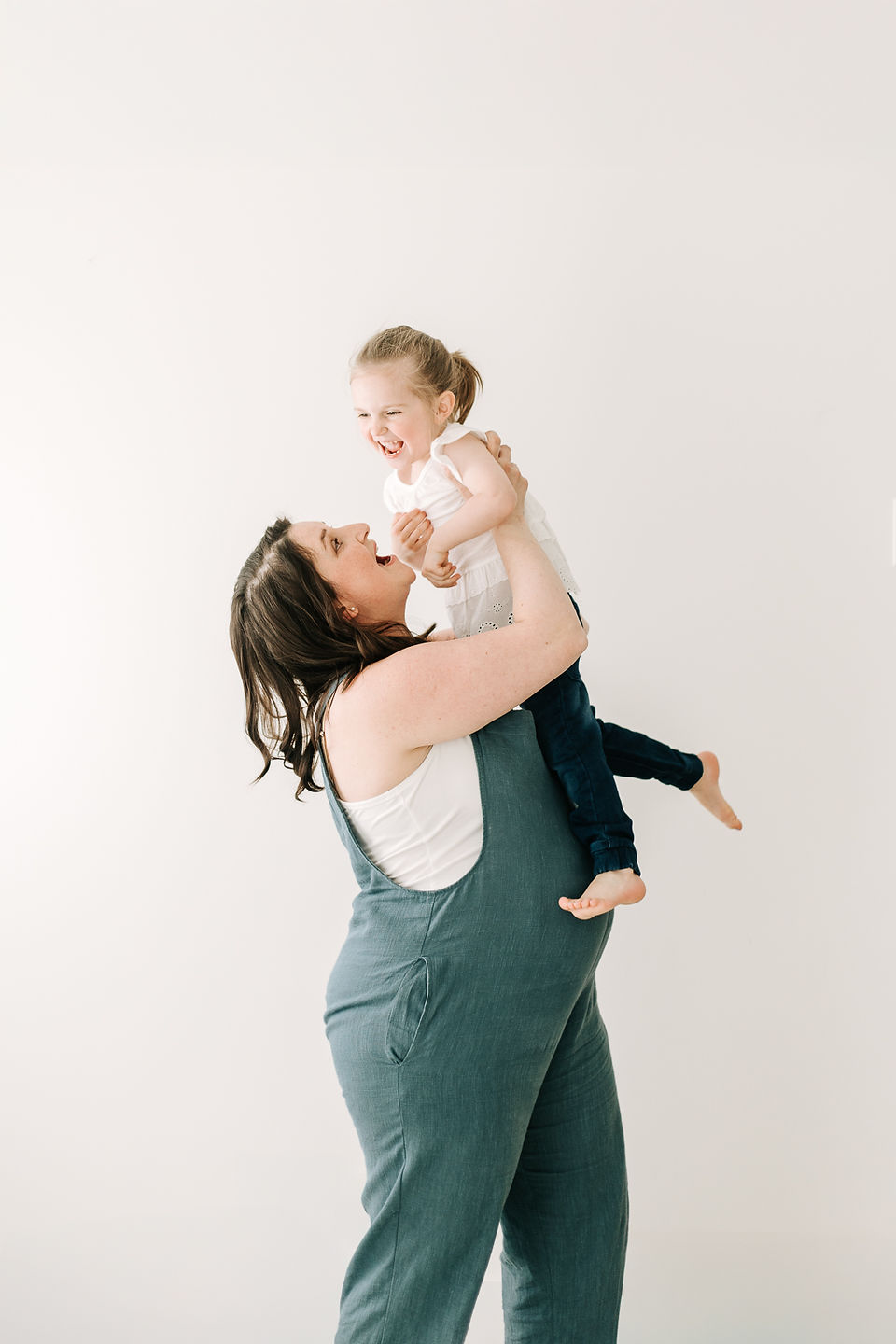 A pregnant woman in blue overalls lifts a young girl in the air as they laugh