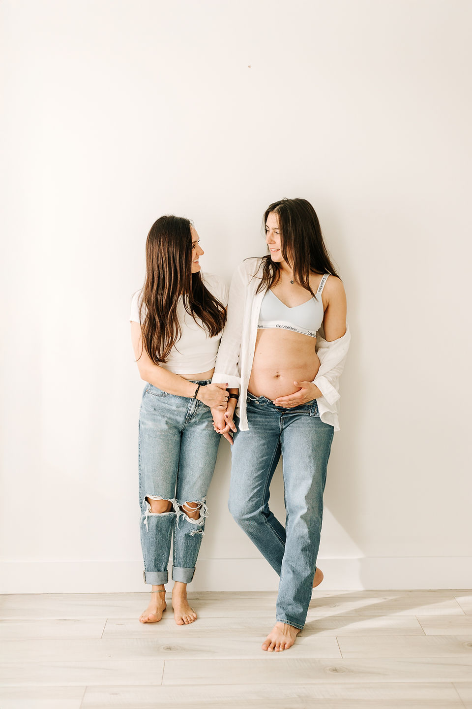 two woman look at each other a hold hands as they lean against a plain wall, one woman in white t shirt and jeans and the other in jeans, a white bra and white button down shirt and pregnant stomach