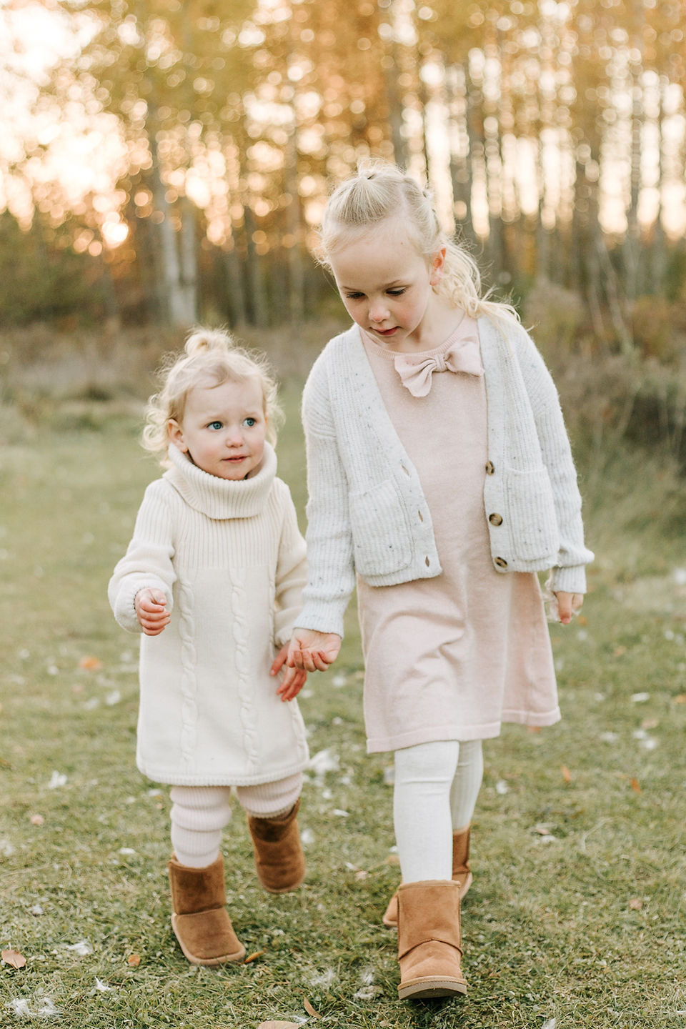 Two girls walk hand in hand on a grass path amongst trees in the fall