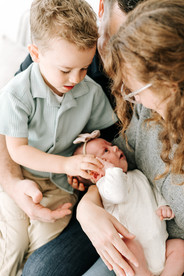 A woman sits on the couch holding a newborn baby while a man sits beside her with a young boy on his lap and the boy holds the baby's hand
