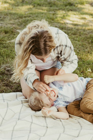 A woman and a young boy play on a blanket in a grassy field under trees