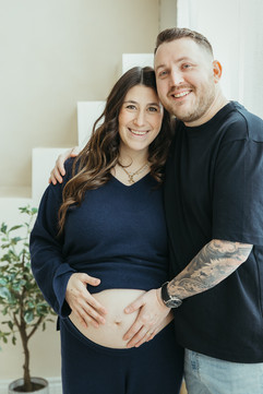 A pregnant woman and a man stand and have their arms around each other as they smile and rest a hand on her pregnant stomach