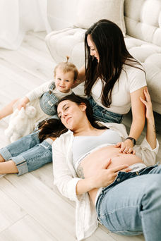 woman in jeans and white t shirt sits on the floor leaning against a couch, while her wife lies on the floor with her head in her lap and both of their hands rest on her pregnant belly. A toddler girl sits beside them