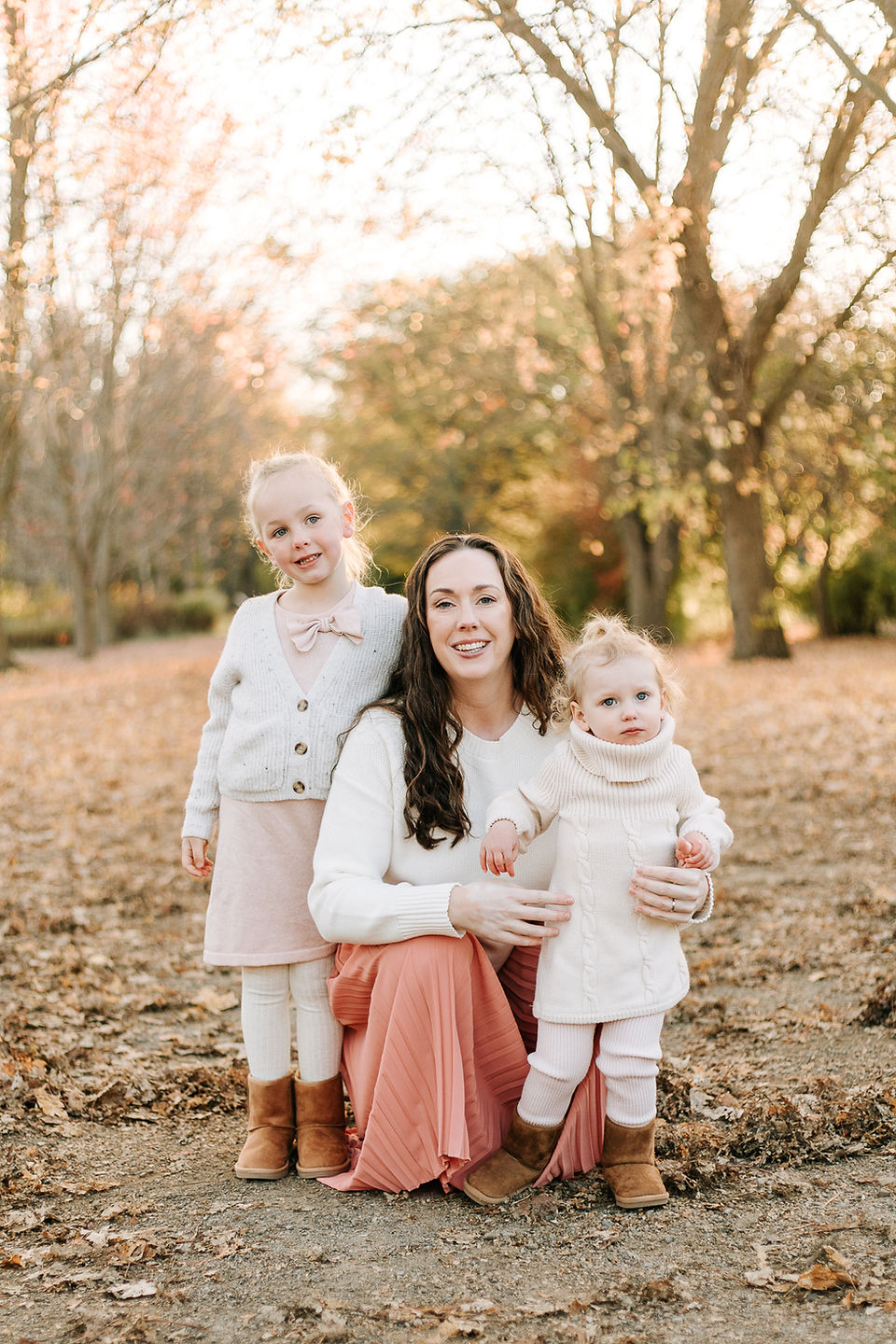 A woman crouches down with two little girls amongst trees in the fall