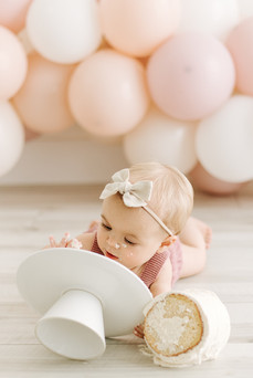 A one year old lies on the floor in front of a balloon arch and eats a small white cake