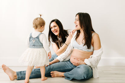 A toddler girl has her back to the camera and is facing her two moms who are sitting on the floor one of whom is pregnant and holding her stomach