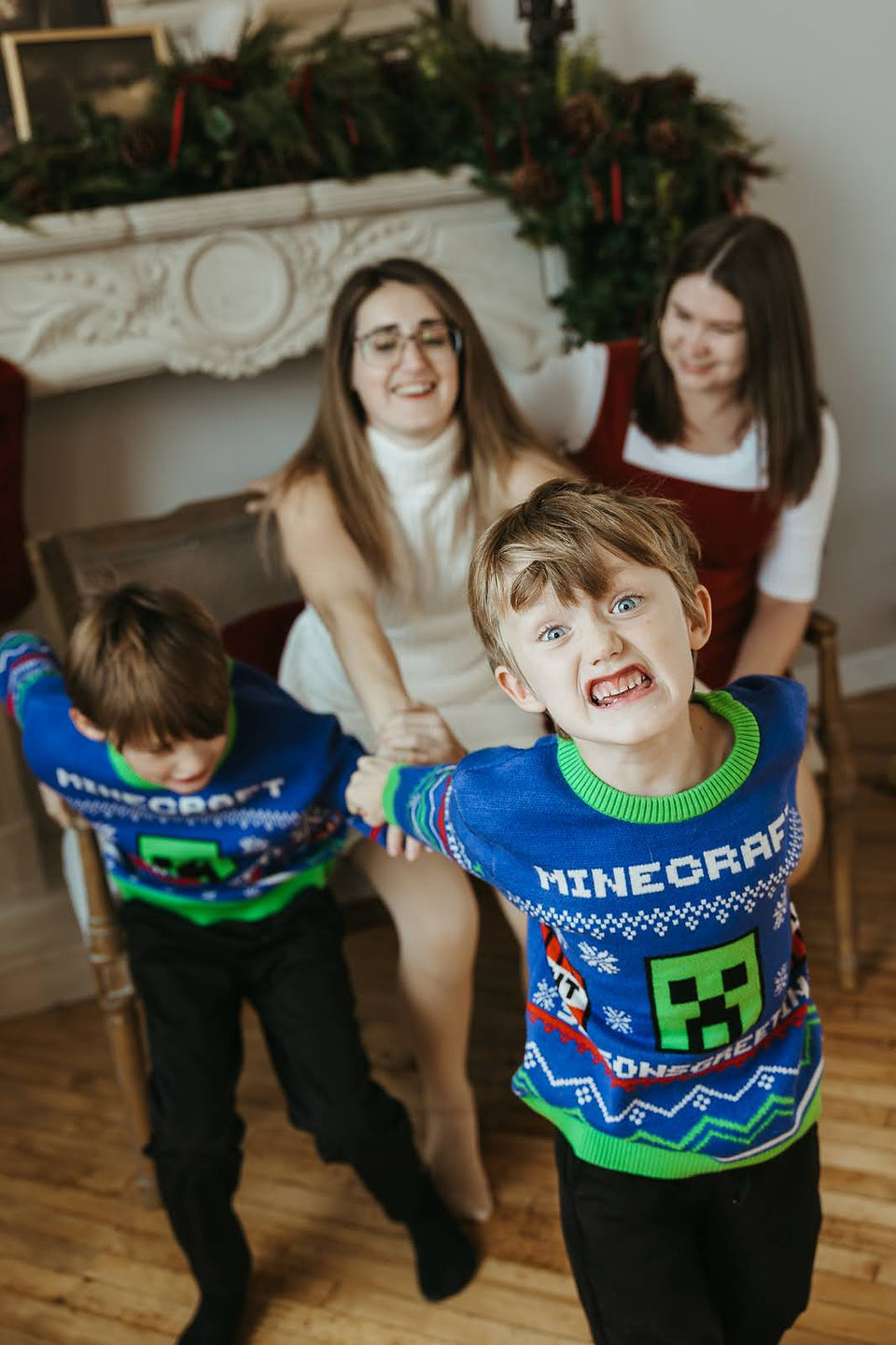 Two woman and two boys play and sit on a couch in front of a fireplace with a Christmas garland on it