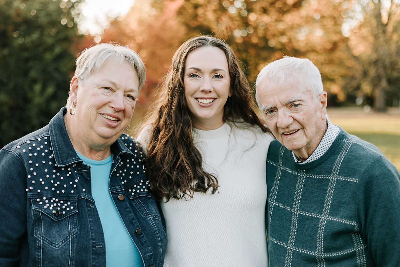 A woman stands between an older man and woman with her arms around them as they all smile