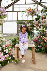 A girl sits on a small wooden bench and smiles as she is surrounded by a large floral arrangement
