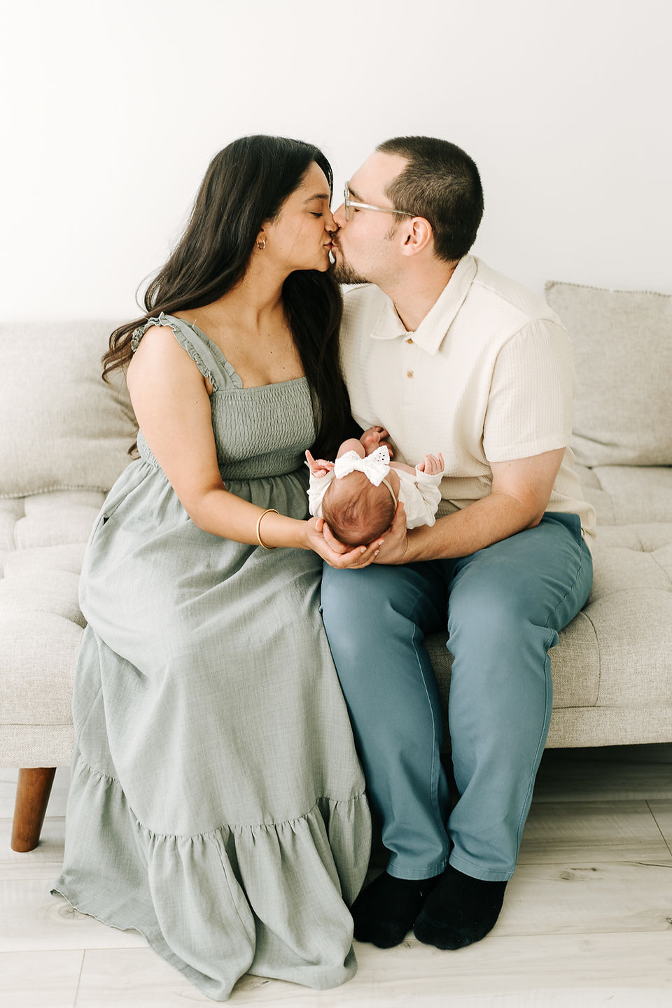 A man and woman sit on a couch and kiss as they hold a newborn baby in their arms 