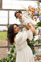 A smiling woman in a light dress lifts a baby in the air in front of a window and floral arrangement