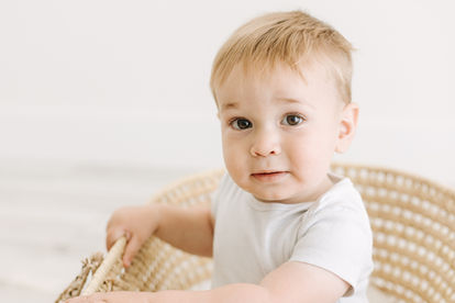 A one year old boy in a white onesie sits in a moses basket