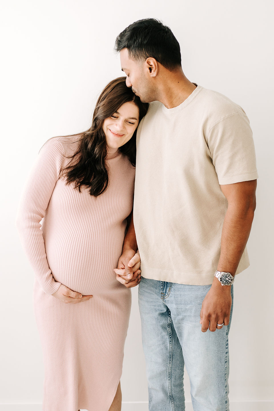woman in a pink dress cradles her pregnant stomach and leans her head against a man's chest while holding his hand and he kisses her head
