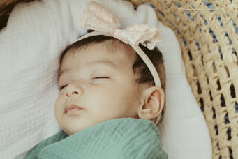 A baby in a pink hair bow and green swaddle sleeps in a bassinet