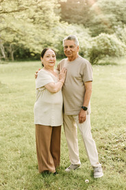 An older couple have their arms around each other and smile as they stand on a grass field 