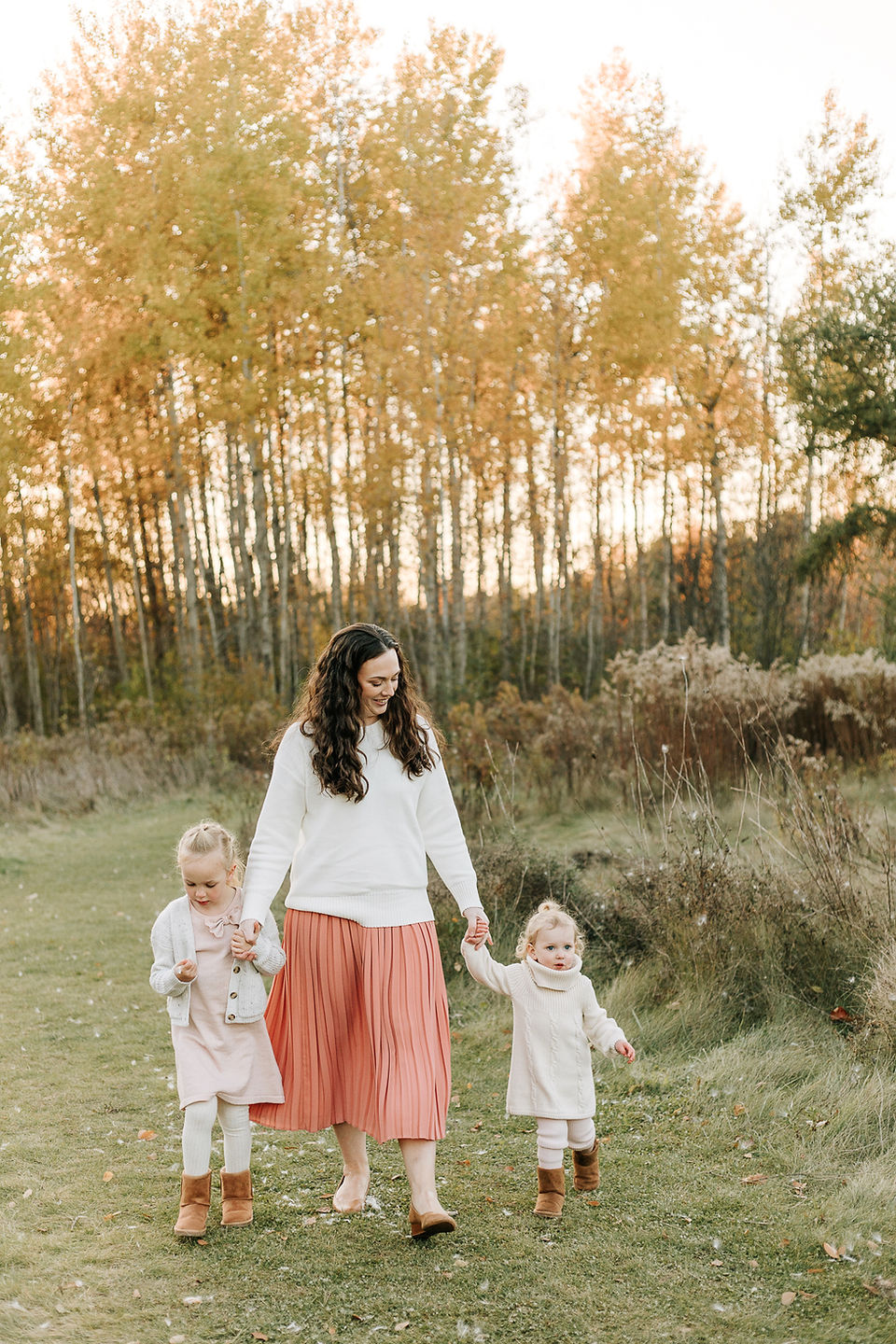 A woman walks hand in hand with two little girls on a grass path amongst trees in the fall