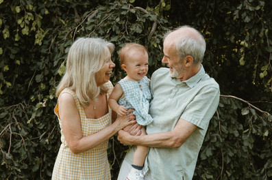 An older man holds a one year old girl and smiles as an older woman stands next to them and rests her hand on the girl as they stand in front of a tree