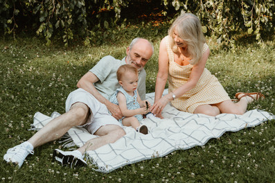 An older man and an older woman sit on a blanket on grass and play with a one year old girl