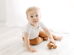 A one year old boy in brown pants and white onesie plays with a small wooden airplane