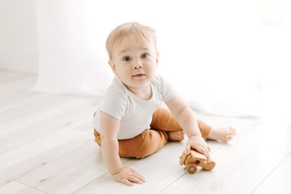 A one year old boy in a white onesie and brown pants plays with a toy on the floor