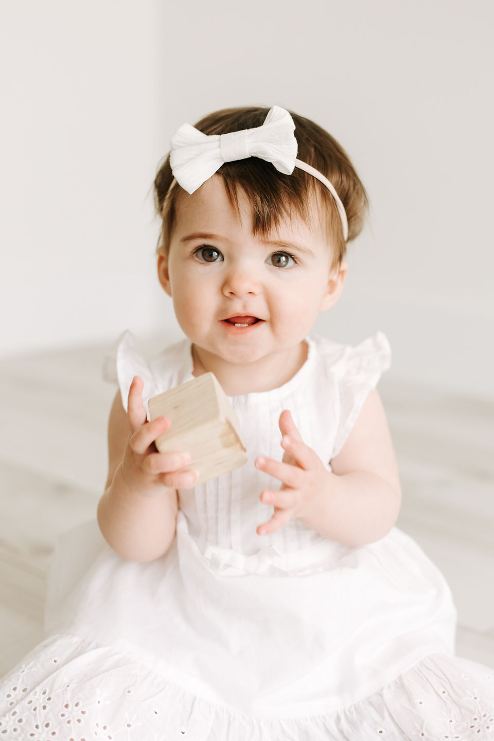 A one year old girl in a white dress sits on the floor and holds a toy