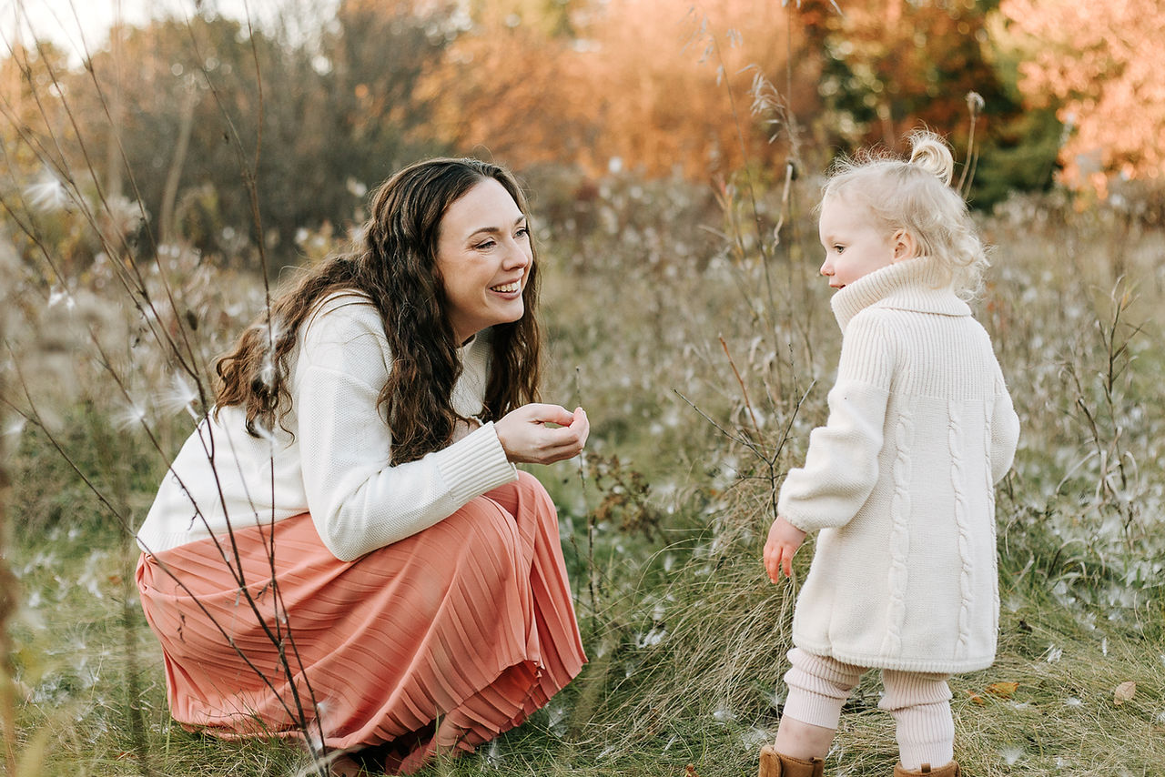 A woman crouches down and smiles at a young girl as they stand in a grass field with milkweed