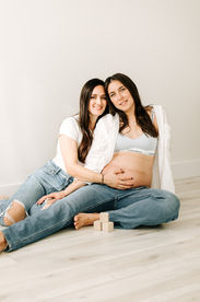 two woman sit on the floor in jeans and white tops, one has her hand on the pregnant stomach of the other