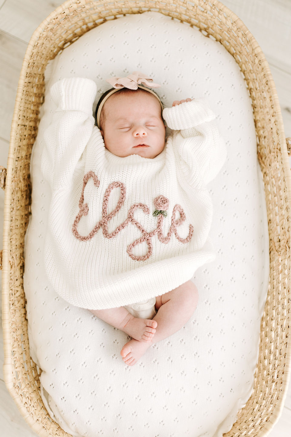 A baby lies in a moses basket wearing a knit sweater that says Elsie