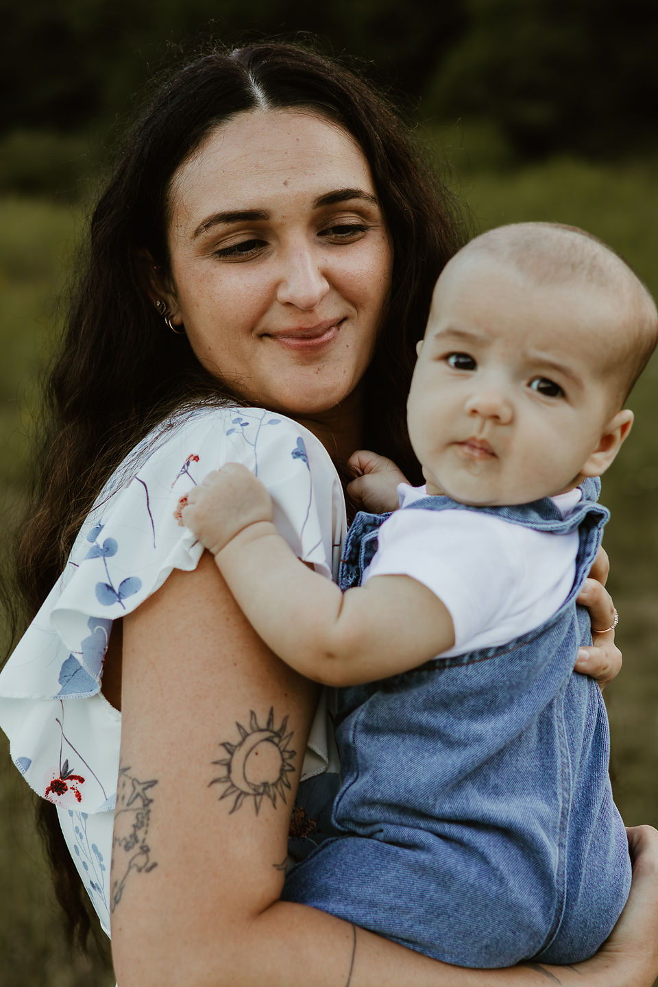 A woman in a white dress with blue flowers on it holds a baby and looks down at him