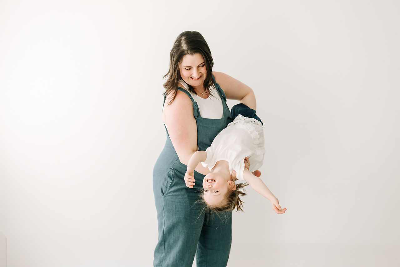 A pregnant woman in blue overalls holds a young girl upside down in her arms as they laugh
