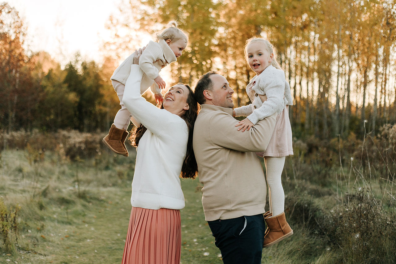 A man and a woman stand back to back as they each lift a young girl in the air on a grassy path under trees in the fall