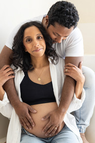A pregnant woman holds onto the arms on a man who sits behind her and looks down and rests his hands on her pregnant stomach