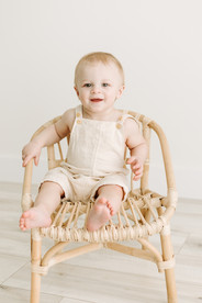 1 year old boy in beige overalls sits on small rattan chair