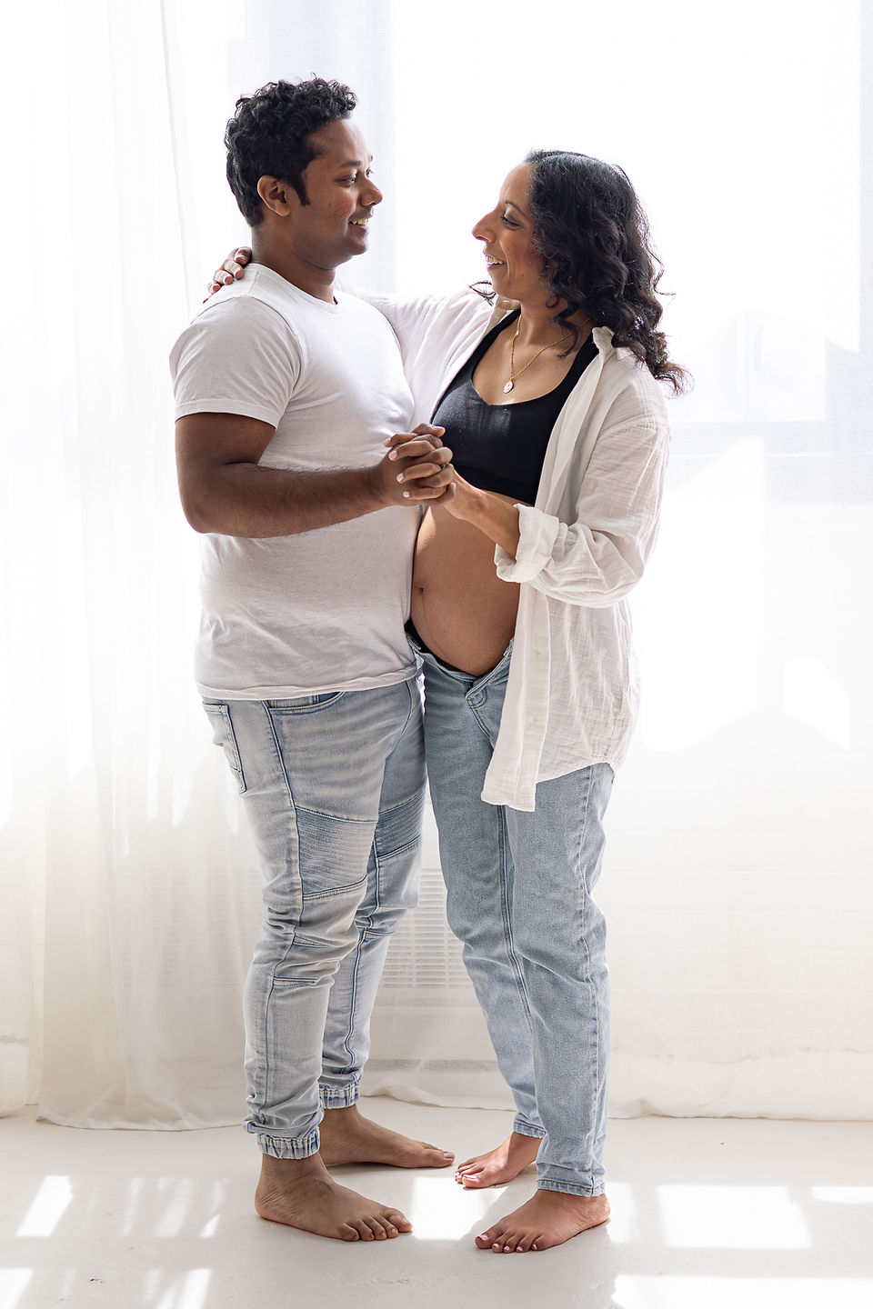 A man in a white t shirt and jeans dances with a pregnant woman in jeans and white shirt in front of a bright window