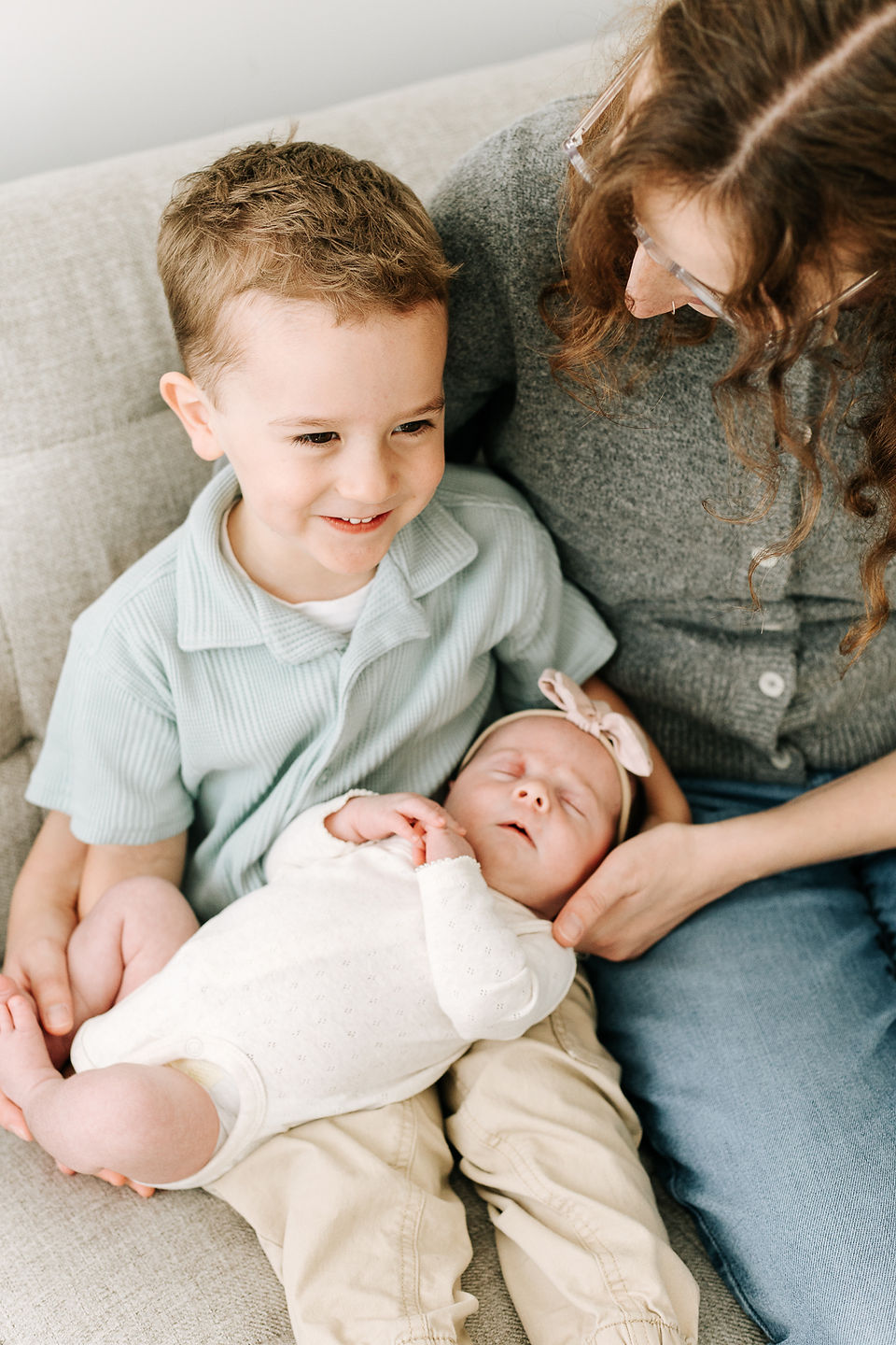 A woman sits on the couch with a young boy and helps him hold a newborn baby