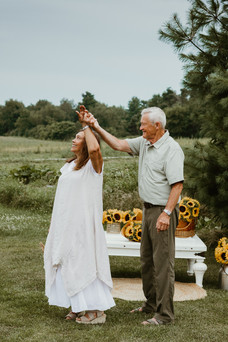 A man and woman dance in a grass field in front of a table with sunflower bouquets on it